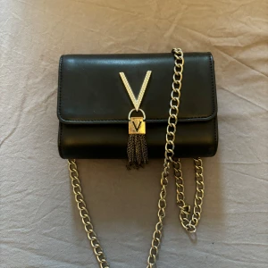 Valentino Svart axelväska med kedja - Snygg svart valentino axelväska i skinn med silverfärgad kedja och dekorativ tofsdetalj. Perfekt för en kväll ute eller en fest. Väskan har en elegant V-formad detalj på framsidan och en praktisk insida för dina nödvändigheter. 