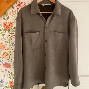 Grå overshirt från Zara - Säljer en stilren grå overshirt från Zara i storlek M. Skjortan har långa ärmar och två praktiska bröstfickor. Perfekt för lager-på-lager under höst och vår. Materialet känns mjukt och bekvämt, och den har knappar framtill för en klassisk look.