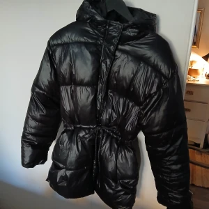 Svart pufferjacka med huva från H&M - Säljer en svart pufferjacka i glansigt material. Jackan har en huva och knyts i midjan för en snygg passform. Perfekt för kalla vinterdagar. Den är i bra skick och redo för nya äventyr!