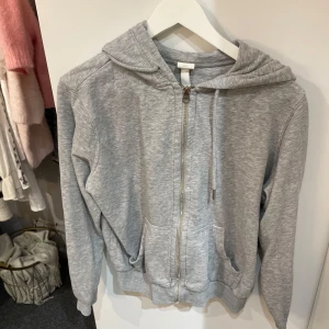 Grå hoodie från H&M - Säljer en grå hoodie från H&M i storlek M men passar även som s. Den har en dragkedja framtill och en mysig huva med snören. Perfekt för en avslappnad stil och passar till alla årstider. Två praktiska fickor framtill för att hålla händerna varma eller förvara småsaker.😍