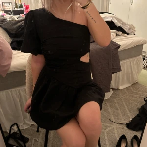 Svart oneshoulder klänning - Snygg svart oneshoulder klänning med en cool cut-out detalj i midjan. Perfekt för festliga tillfällen! Den har en kort längd och en voluminös kjol som ger en fin siluett. Materialet ser ut att vara mjukt och bekvämt.