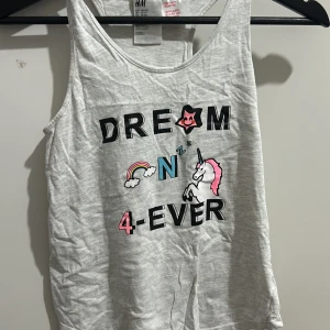 Grå linne från H&M med enhörningstryck - Säljer ett grått linne från H&M med ett coolt tryck av en enhörning och texten 'Dream On 4-Ever'. Perfekt för sommarens alla äventyr! Linnet är i bomull och har en avslappnad passform. Superfint och bekvämt för varma dagar. 🦄✨