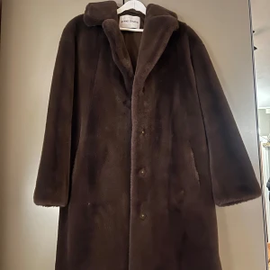 Brun pälsjacka från Stand Studio - Säljer en supermjuk och lyxig brun pälsjacka från Stand Studio. Den har en klassisk design med långa ärmar och knappar framtill. Perfekt för att hålla sig varm och stilren under kalla dagar. Passar både till vardags och festliga tillfällen!