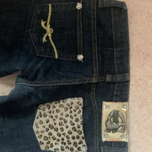 Mörkblå jeans med leoparddetaljer - Säljer ett par mörkblå jeans med coola leopardmönstrade fickdetaljer och glittriga knappar. De har en tight passform och låg midja. De är äckta just cavalli. Storleken på lappen är 27. Midjemåttet är 37cm. Innerbenslängden är 72cm. Observera att hela jeansen är glittriga. Kan sänka priset vid snabbt köp! 