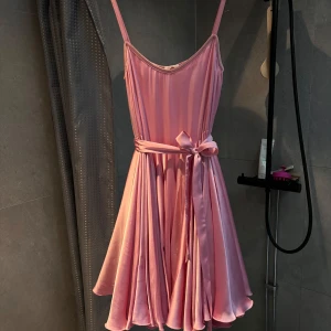 Rosa Ida Sjöstedt-klänning - Superfin rosa klänning i satin med ett knytband i midjan. Axelband och en härlig, fladdrig kjol som passar perfekt för sommarens fester. Klänningen är i mycket bra skick och ger en söt och elegant look.