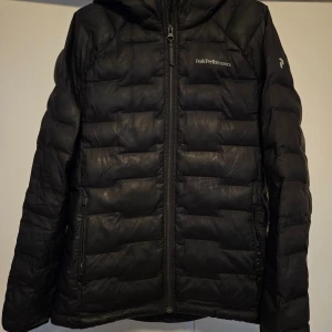 Svart dunjacka från Peak Performance - Säljer en svart dunjacka från Peak Performance i mycket bra skick. Jackan är perfekt för kalla vinterdagar med sin quiltade design och långa ärmar. Den har en praktisk dragkedja framtill och en skön huva. Passar både till vardags och utomhusaktiviteter. Storleken är inte specificerad, men den ser ut att vara normal passform.