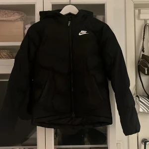 Svart pufferjacka från Nike - Säljer en svart pufferjacka från Nike i storlek M. Jackan har en dragkedja framtill och en huva för extra skydd mot väder och vind. Perfekt för kyliga dagar och har en stilren design med Nike-loggan på bröstet. Passar både till vardags och sportiga aktiviteter.