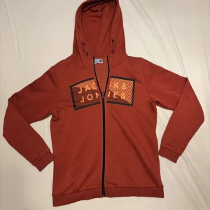 Rostfärgad hoodie från Jack & Jones - Säljer en snygg rostfärgad hoodie från Jack & Jones. Den har en dragkedja framtill och ett coolt tryck på bröstet. Perfekt för höst och vår när du vill hålla det casual men ändå stiligt. Passar bra till jeans eller joggers!