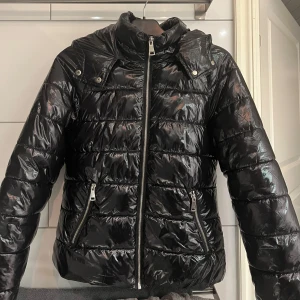 Svart pufferjacka från Nelly - Säljer denna populära och snygga svarta pufferjackan från Nelly i storlek 38. Jackan har en glansig finish med dragkedja framtill och två  fickor med dragkedjor. Perfekt för kyliga dagar nu till vinter. Använd ett fåtal gånger därmed är som ny. nypris 800kr 
