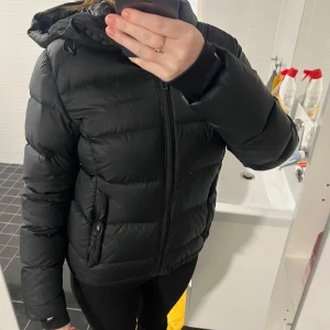 Svart pufferjacka med huva - Säljer en snygg svart pufferjacka i mycket bra skick. Jackan har en dragkedja framtill och en praktisk huva. Perfekt för kalla dagar och håller dig varm och bekväm. Passar till både vardag och utflykter. Storlek dam M.