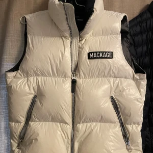 Beige dunväst från Mackage - Säljer en snygg beige dunväst från Mackage i mycket bra skick. Västen har en quiltad design med dragkedja framtill och två praktiska fickor med dragkedjor. Perfekt för höst och vår när du vill hålla dig varm och stilren. Stor logga på bröstet ger en cool touch.