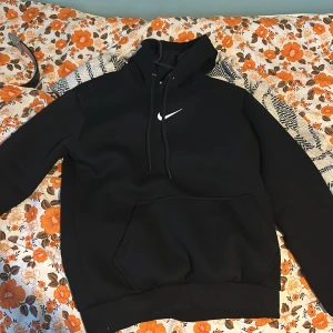 Svart hoodie från Nike - Säljer en klassisk svart hoodie från Nike i storlek S. Den har en vit Nike-logga på bröstet och en praktisk magficka. Perfekt för både vardag och träning. Huvan har dragsko för justerbar passform. Superbekväm och stilren och väldigt varm