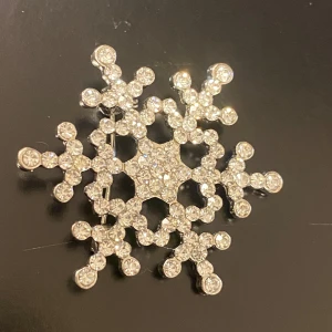 Snöflingeformad brosch med strass - En glittrig brosch i form av en snöflinga, täckt med gnistrande strass. Perfekt för att ge en vinterkänsla till din outfit. Den är i silverfärg och passar utmärkt till festliga tillfällen eller som en fin detalj på en kappa eller tröja.