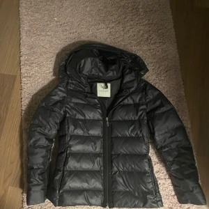 Svart dunjacka från Moncler - Säljer en snygg svart dunjacka från Moncler i mycket bra skick. Jackan har en stilren design med dragkedja och huva, perfekt för kyliga dagar. Den är både varm och bekväm, idealisk för höst och vinter. Passar perfekt för dig som vill ha både stil och funktion. Jackan är en gammal modell och saknar där av serie tidning designen 