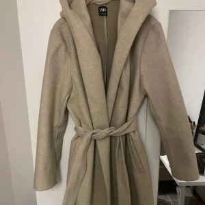Beige kappa från Zara - Säljer en superfin beige kappa från Zara i storlek M men passar även S då jag har st S vanligtvis . Den har en snygg knytning i midjan och en stor huva för extra mysfaktor. Perfekt för höst och vår! Kappan är i mycket bra skick och gjord av ett mjukt material som känns lyxigt. Passar både till vardags och fest. 🌟