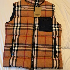 Rutig dunväst från Burberry - Säljer en snygg rutig dunväst från Burberry i klassiskt mönster. Västen är vändbar med en sida i marinblått och den andra i rutigt mönster. Den har dragkedja och en praktisk ficka på bröstet. Perfekt för höst och vår! Ps. Oanvänd 