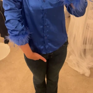 Blå satinblus med fjäderdetaljer - Säljer en supercool blå satinblus med fjäderdetaljer vid ärmsluten. Blusen har knappar framtill och en glansig finish som gör den perfekt för festliga tillfällen. Den är långärmad och ger en lyxig känsla. Passar perfekt till både jeans och kjol för en chic look!