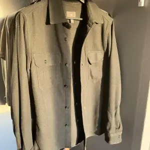 Säljer en stilren svart overshirt från H&M i mycket bra skick. Den har långa ärmar och knappar framtill samt två bröstfickor med lock. Perfekt för höst och vår, kan bäras som jacka eller skjorta. Passar både till vardags och fest!