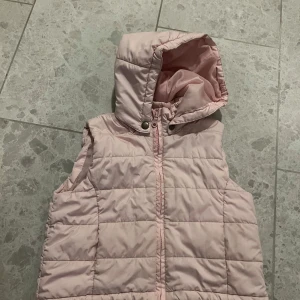Rosa dunväst från Kids-Up - Säljer en superfin rosa dunväst från Kids-Up i mycket bra skick. Västen har en praktisk huva och dragkedja framtill. Perfekt för kyliga dagar när man vill hålla sig varm och snygg. Passar perfekt för vår och höst!