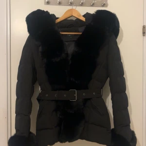 Svart dunjacka med pälsdetaljer - Säljer en snygg svart dunjacka i helt nytt skick. Jackan har en fluffig pälsdetalj runt huvan och längs med dragkedjan. Den har ett bälte i midjan för en figursydd look och långa ärmar. Perfekt för kalla vinterdagar. Jag köpte jackan för 3400kr men säljer den för 2500. 