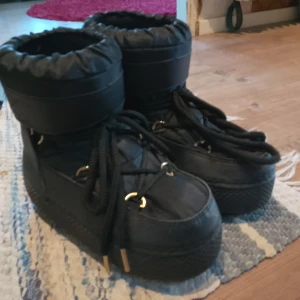 Vinter skor - Säljer ett par svarta moon boots i syntetmaterial. De har en hög och vadderad design med snörning framtill. Perfekta för vinterns kalla dagar och håller fötterna varma och torra. Använda men i bra skick. Passar perfekt för snöiga äventyr! ❄️