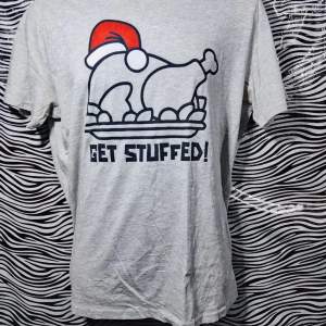 Säljer en grå t-shirt med ett roligt julmotiv av en kalkon med tomteluva och texten 'GET STUFFED!'. Perfekt för julfesten eller som en kul present. T-shirten är kortärmad och i bomull, vilket gör den bekväm att bära hela dagen.