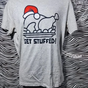 Grå t-shirt med julmotiv - Säljer en grå t-shirt med ett roligt julmotiv av en kalkon med tomteluva och texten 'GET STUFFED!'. Perfekt för julfesten eller som en kul present. T-shirten är kortärmad och i bomull, vilket gör den bekväm att bära hela dagen.