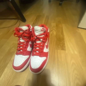 Nike Dunk i rött och vitt - Säljer ett par Nike Dunk i rött och vitt. Skorna är i mycket bra skick och har en klassisk design med snörning. Perfekta för både vardag och fest! De har en snygg kontrast mellan det röda och vita, vilket gör dem riktigt iögonfallande.