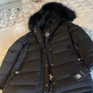 Svart dunjacka från Hollies - Säljer en svart dunjacka från Hollies i mycket bra skick. Jackan har en fluffig pälsfodrad huva och stängs med dragkedja och knappar. Perfekt för kalla vinterdagar med sin varma och stilrena design. Den har långa ärmar och praktiska fickor med dragkedja. Passar perfekt för vinterns alla äventyr!  Fick den u present men de va lite för stor så bestämde mig för att sälja den! Jackan kostar 5000kr säljs för 2500kr