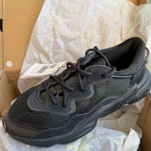 Svarta Adidas Ozweego sneakers - Säljer ett par svarta Adidas Ozweego sneakers i nytt skick. De har en cool och chunky design med snörning och är gjorda i syntetmaterial. Perfekta för både vardag och träning. Skorna kommer i originalkartong.