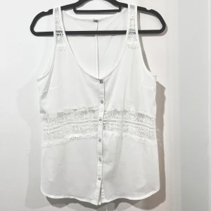 Vit ärmlös blus med spetsdetaljer - Säljer en elegant vit blus med spetsdetaljer och knappar framtill. Den är ärmlös och har en v-ringad design som ger en stilren look. Genomskinlig vid spetspartier. 