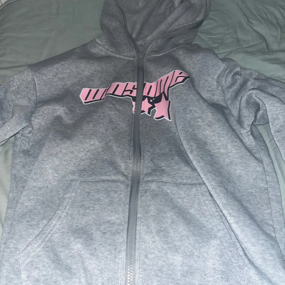 Säljer en grå zip hoodie med dragkedja och rosa stjärndetaljer. Den har en stor rosa stjärna på ärmen och ett coolt tryck på framsidan. Perfekt för en avslappnad stil och mysiga dagar. Hör av dig om du är intresserad! 💫. Hoodies.