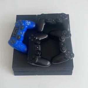PlayStation 4 med fyra kontroller - Säljer en PlayStation 4 med tre svarta och en blå kontroll. Den blå kontrollen har ett unikt mönster och logotyp. Konsolen är i bra skick och perfekt för spelkvällar med vänner. Alla kontroller är trådlösa och fungerar utmärkt. Perfekt för den som vill ha en komplett spelupplevelse! 🎮