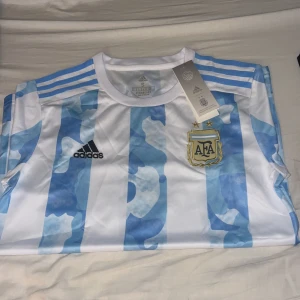 Vit och blå fotbollströja från Adidas - Säljer en snygg vit och blå randig fotbollströja från Adidas med AFA-logga på bröstet. Tröjan är i nytt skick och har korta ärmar. Perfekt för fotbollsfans som vill visa sitt stöd. Tillverkad med AEROREADY-material för att hålla dig sval och bekväm.