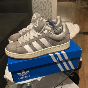 Grå Adidas Campus sneakers - Säljer ett par snygga grå Adidas Campus sneakers i mycket bra skick. De har en klassisk design med vita ränder och snörning. Perfekta för både vardag och fest. Skorna är i storlek herr 42 och kommer i originalkartong.