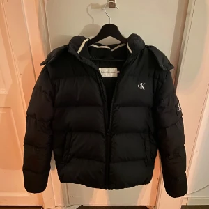 Svart pufferjacka från Calvin Klein - Hej, jag säljer en stilren pufferjacka från Calvin Klein i storlek M. Jackan är väldigt bekväm och perfekt för kalla vinter dagar. Jackan är i gott skick men med några få defekter. Jackan har en klassisk design med CK-loggan på bröstet. En perfekt jacka för vintern! ❄️☃️