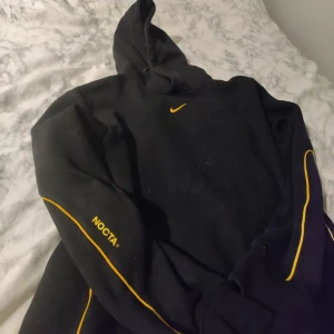 Svart hoodie från NOCTA - Säljer en svart hoodie från NOCTA med gula detaljer och logga. Den har en klassisk passform med huva och långa ärmar. Perfekt för höst och vinter, både stilren och bekväm. Passar lika bra till vardags som till träning.