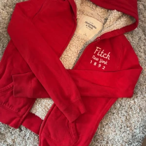 Röd hoodie från Abercrombie & Fitch - Säljer en snygg röd hoodie från Abercrombie & Fitch med vit broderad text 'Fitch New York 1892'. Den har en mysig fleecefodring och dragkedja framtill. 💝