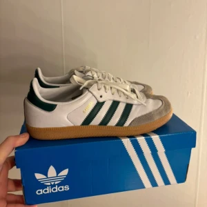 Adidas Samba OG i vitt och grönt - Säljer ett par klassiska Adidas Samba OG sneakers i vitt och grönt. Skorna har en snygg design med gröna ränder och en gummisula. Perfekta för både vardag och träning. De är i nytt skick och kommer i originalkartong.