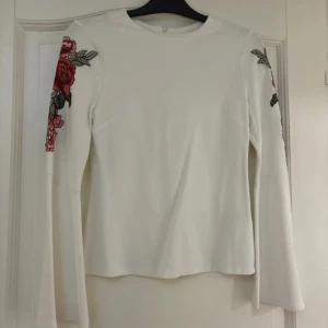 Vit långärmad tröja med blombroderi - Säljer en superfin vit tröja med långa ärmar och vackra blombroderier i rött och grönt på ärmarna. Tröjan har en dragkedja i ryggen för enkel på- och avtagning. Knappt använd, därav mycket bra skick