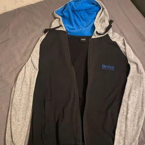 Svart och grå hoodie från Boss - Säljer en snygg hoodie från Boss i svart och grått med blått foder i luvan. Den har dragkedja framtill och är långärmad. Perfekt för höst och vår, och passar både till vardags och träning. Boss-loggan är broderad i blått på bröstet. Skön och stilren design!