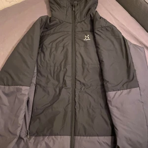 Svart och grå jacka från Haglöfs - Säljer en snygg svart och grå jacka från Haglöfs. Den har en praktisk dragkedja och huva, perfekt för kyliga dagar. Jackan är i mycket bra skick och passar både till vardags och utomhusaktiviteter. Perfekt för höst och vår!