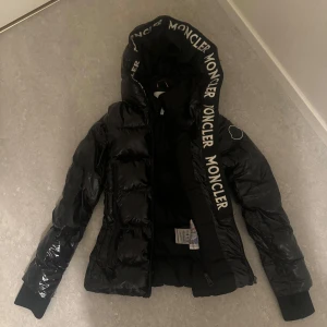 Svart dunjacka från Moncler - Säljer en snygg svart jacka med stor Moncler logga på huvan. Jackan har en glansig finish och dragkedja framtill. Perfekt för kyliga dagar, säljer pågrund av att den inte kommer till nån användning längre
