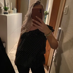 Svart prickig oneshoulder topp - Säljer en superfin svart oneshoulder topp med vita prickar. Den har en volangdetalj som ger en extra touch av stil. Perfekt för sommarens fester eller en utekväll. Toppen är i bra skick och passar perfekt till både jeans och kjol.