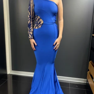 Blå one-shoulder klänning - Snygg blå långklänning med one-shoulder design och en ärm med spetsdetaljer. Perfekt för festliga tillfällen med sin eleganta passform och unika stil. Klänningen har en smickrande siluett och är tillverkad i ett mjukt material som ger en bekväm känsla.