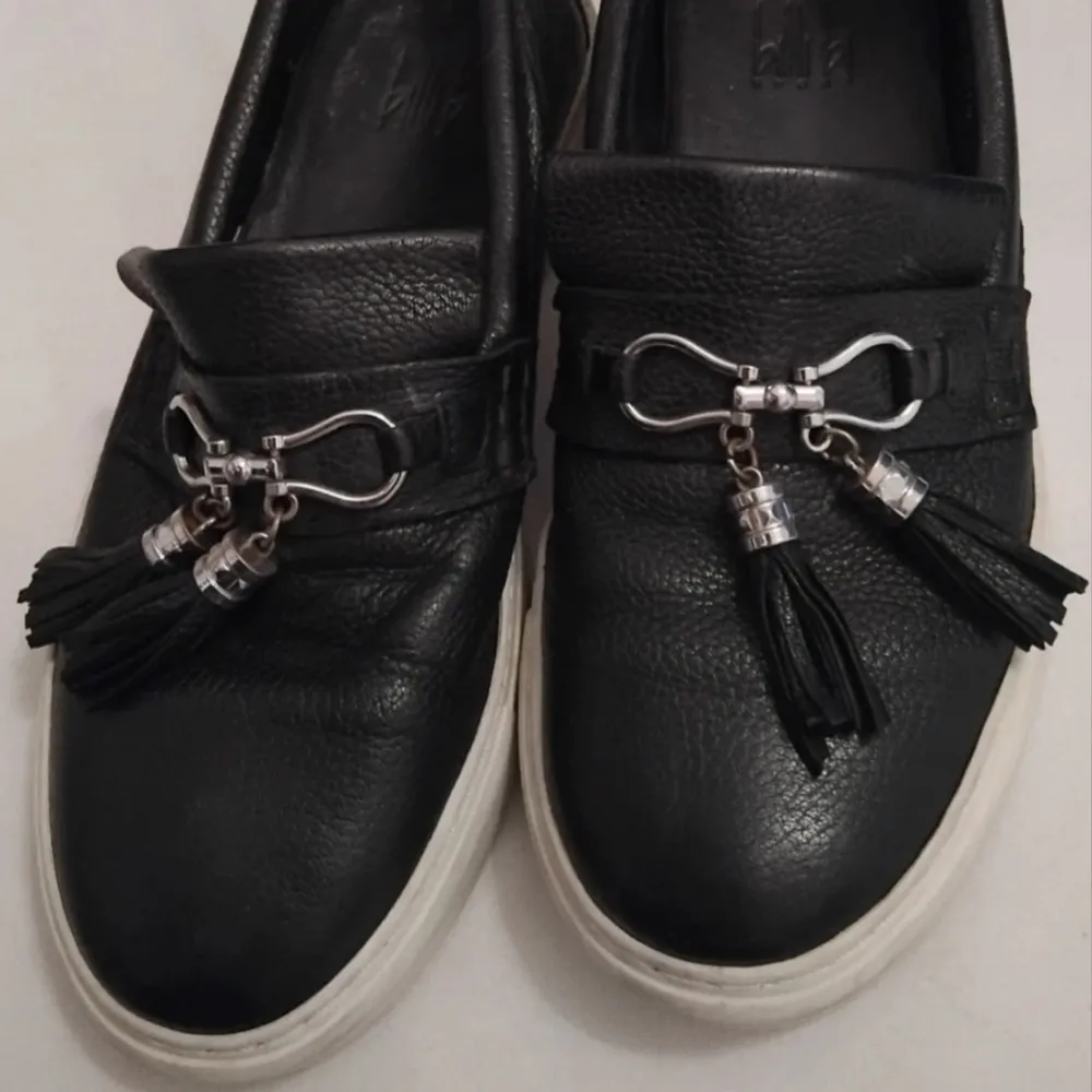 Snygga svarta loafers i skinn med coola tofsdetaljer och silverfärgad dekoration framtill. De har en vit sula som ger en skön kontrast och en stilren look. Perfekta för både vardag och festliga tillfällen. Är lite missfärgning på det vita men kanske kan skrubbas bort, annars I fint skick. Dyra I inköp.. Skor.