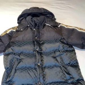 Gucci puffer - Säljer en snygg svart pufferjacka från Gucci med deras ikoniska mönster. Jackan har långa ärmar och en huva för extra värme. Den är dekorerad med en beige rand med logotyp på ärmarna. Perfekt för vintern och hösten!