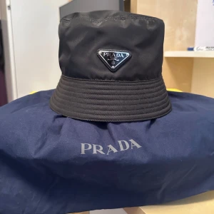 Svart bucket hat från Prada - Snygg svart bucket hat från Prada i ett stilrent och modernt utförande. Hatten har en broderad logga framtill och är perfekt för både vardag och fest. Materialet känns lyxigt och hållbart. Kommer med en blå förvaringspåse med Prada-logga. 