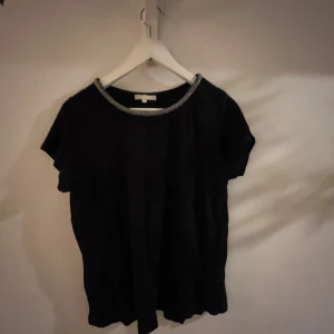 Svart topp med glittrig halsringning - Snygg svart topp från Maje med en glittrig halsringning som ger en elegant touch. Perfekt för att lyfta vilken outfit som helst. Kortärmad och i en bekväm passform.