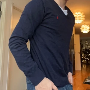 Mörkblå tröja från Ralph Lauren - Säljer en stilren mörkblå tröja från Ralph Lauren med v-ringning och ett litet rött broderat märke på bröstet. Tröjan har långa ärmar och är perfekt för en avslappnad men ändå elegant look.
